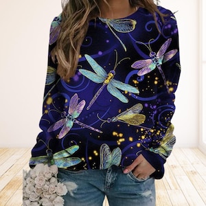 Può includere: Una maglia a maniche lunghe con sfondo blu scuro e un motivo di libellule colorate. Le libellule sono in tonalità di blu, viola e giallo, con alcuni piccoli punti gialli sparsi.