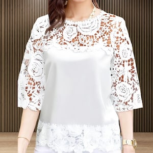 May include: White blouse with a floral lace overlay on the sleeves and upper chest. The blouse has a round neckline and three-quarter sleeves. The bottom hem is also trimmed with lace. The blouse is made of a soft, flowing fabric.