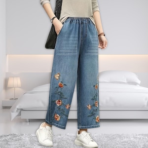 Könnte beinhalten: Hellblaue Jeans-Hose mit weitem Bein und elastischem Bund. Die Hose hat gestickte Blumen- und Schmetterlingsdesigns in Orange-, Rot- und Grüntönen. Das Model trägt weiße Turnschuhe.