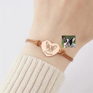 Könnte beinhalten: Ein roségoldfarbenes Armband mit herzförmigem Anhänger, das eine gravierte Strichzeichnung einer Französischen Bulldogge und den Namen Charlie zeigt. Der Anhänger ist an einem dünnen, hellbraunen Band befestigt und zeigt ein individuelles Haustierporträt.