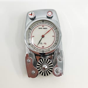 May include: A chrome-finished Tape Timer device featuring a circular analog clock face with red hands, black numerals, and the text TAPE TIMER. The unit includes two top adjustment knobs and a central black and white striped circular dial.