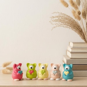 Könnte beinhalten: Fünf kleine, stehende Monsterfiguren in einer Reihe, jede mit einem großen zentralen Auge und einem kleinen Herz in den Händen. Die Figuren sind rosa, hellgrün, gelb, pfirsichfarben und blau, alle mit weißen Füßen und schwarzen Pupillen.