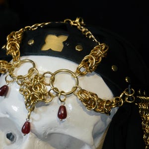 Puede incluir: Un tocado de cota de malla en tono dorado con anillos metálicos entrelazados y cuentas colgantes de color rojo oscuro en forma de lágrima. El tocado está sujeto a una banda de cuero negro con remaches dorados y un detalle recortado.