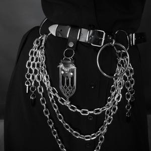 May include: Black leather belt with silver metal buckle and chain detailing. The belt has a decorative bat-shaped pendant and multiple hanging chains. A large silver ring is attached, enhancing the gothic style of the belt.