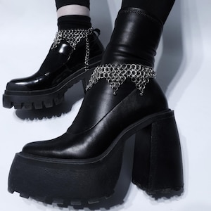 May include: Black platform boots with chunky soles and block heels. The boots have a glossy finish and are adorned with silver chain anklets. The boots are worn with black socks and are set against a white background.