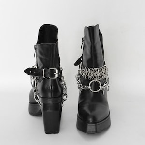 Puede incluir: Botas de tobillo de cuero negro con detalles de cadena y hebilla plateadas. Las botas tienen un tacón grueso y una punta puntiaguda.