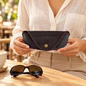 May include: A dark brown leather glasses case with a curved flap, secured by a single brass-colored metal snap button. The edges of the case are finished with a decorative dark brown whipstitch detail. A pair of dark-rimmed sunglasses sits nearby.