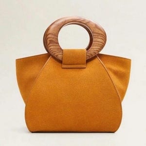 May include: Mustard-colored handbag with a wooden handle. The bag has a rounded, trapezoid shape with a suede-like texture. The handle is a large, circular wooden ring attached to the top of the bag with a small flap.