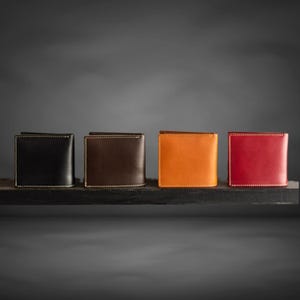 Op de afbeelding: Vier leren portemonnees in verschillende kleuren: zwart, bruin, oranje en rood. De portemonnees zijn in een rij op een donker oppervlak gerangschikt.