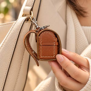 May include: A miniature brown leather backpack charm featuring white contrast stitching, a structured frame, and a silver-toned metal lobster clasp. The small accessory is designed to attach to larger bags, showcasing a detailed, compact construction.
