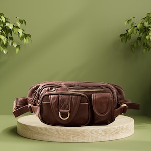 Puede incluir: Un bolso de cintura de cuero marrón con múltiples compartimentos y bolsillos con cremallera. La bolsa presenta una hebilla y cremalleras doradas. El bolso de cintura se exhibe sobre un soporte de madera de color claro sobre un fondo verde.