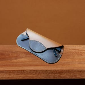 May include: A brown leather glasses case with a flap that folds over to protect the glasses. The case has a dark brown leather lining and stitching around the edges.