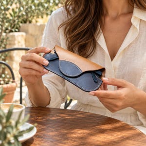 May include: A two-tone eyeglass case featuring a tan leather top flap and a dark navy blue leather base. The case is secured with a rounded flap closure and is accented with visible white stitching along the edges of the navy blue section.