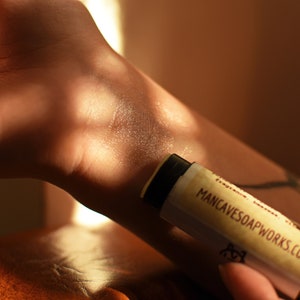 May include: A close-up of a person's arm with a beige lip balm tube being applied. The tube is labeled "MANCAVESOAPWORKS.COM".