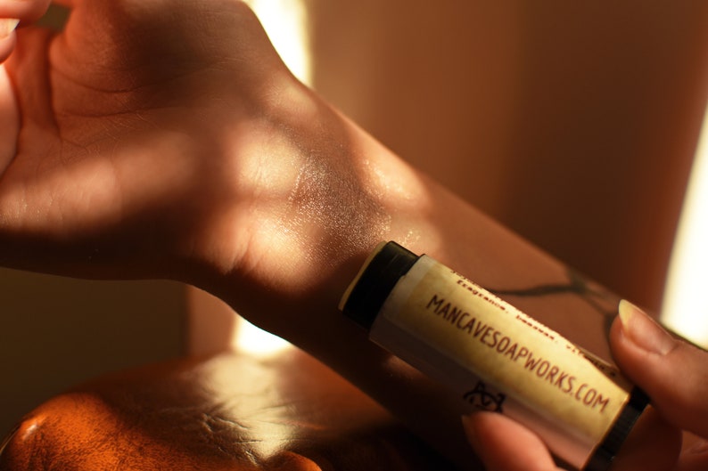 May include: A close-up of a person's arm with a beige lip balm tube being applied. The tube is labeled "MANCAVESOAPWORKS.COM".