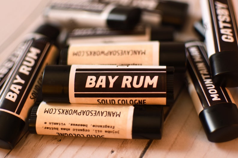 May include: A close-up of a black and white label on a solid cologne stick. The label reads "Bay Rum Solid Cologne" and "MancaveSoapWorks.com".