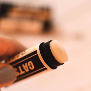 May include: A close-up of a black and white lip balm tube with the word "GATS" printed on the side. The balm is a light beige color.