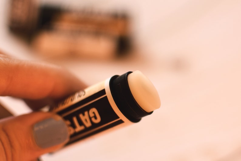 May include: A close-up of a black and white lip balm tube with the word "GATS" printed on the side. The balm is a light beige color.