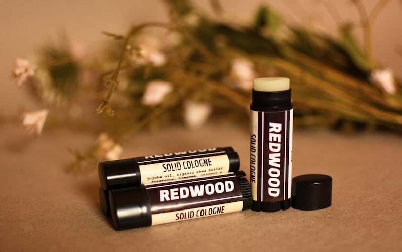 May include: Three black tubes of Redwood solid cologne. The tubes are labeled with the brand name and the words "Solid Cologne". The tubes are on a brown surface.