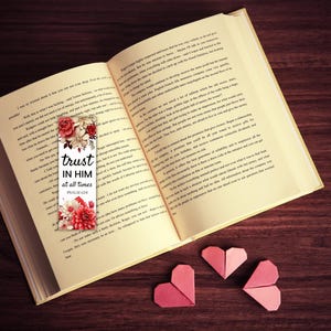 May include: An open book with a floral bookmark that reads "trust in him at all times." The bookmark features red and white flowers. Three origami hearts in shades of pink are arranged near the book on a dark wood surface.