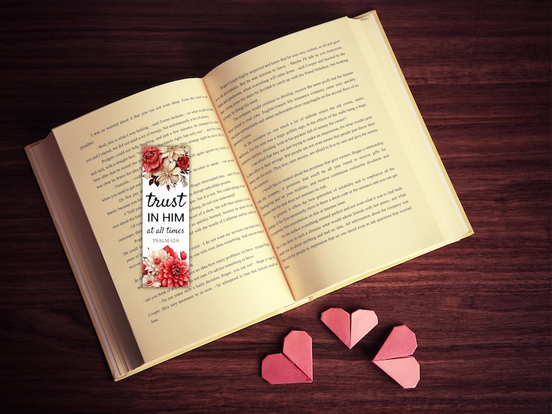 May include: An open book with a floral bookmark that reads "trust in him at all times." The bookmark features red and white flowers. Three origami hearts in shades of pink are arranged near the book on a dark wood surface.