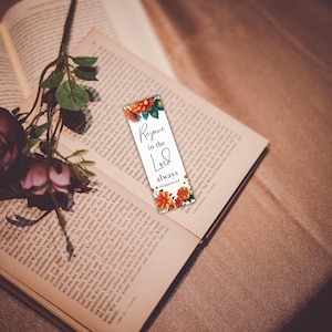 May include: A bookmark with the text "Rejoice in the Lord always" and floral accents rests on an open book. A dark pink rose and greenery are placed on the book. The bookmark is white with orange and green floral designs.