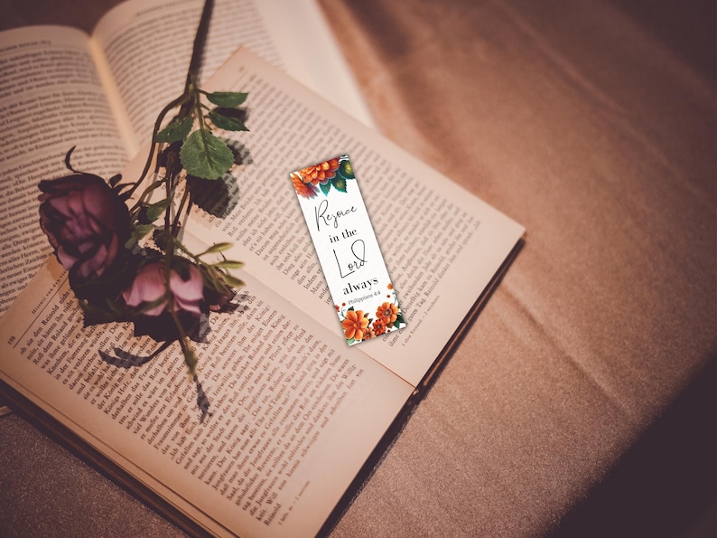 May include: A bookmark with the text "Rejoice in the Lord always" and floral accents rests on an open book. A dark pink rose and greenery are placed on the book. The bookmark is white with orange and green floral designs.
