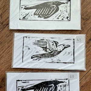 May include: Three rectangular white cards featuring black ink block prints of birds, each enclosed in a clear plastic sleeve. Two cards display a bird in profile, while the center card shows a bird in flight. Each card has a small label marked £3.