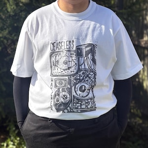 May include: White t-shirt with a black graphic print. The design includes the text "CEASELESS WATCHER" and stylized cameras and eyes. The shirt has a crew neck and short sleeves.