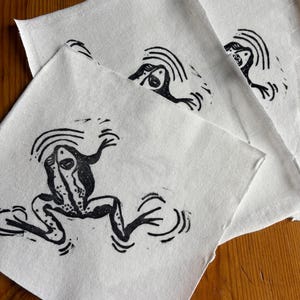 May include: Three white fabric squares feature a black frog linocut print. The frog is depicted with outstretched limbs and a dotted pattern. The linocut style gives a handmade, artistic feel to the fabric.