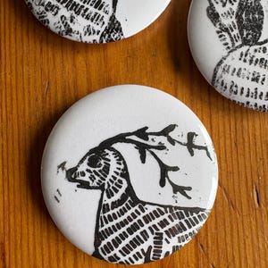 May include: Round white ceramic buttons featuring black deer designs. The deer are depicted in a geometric style with antlers. The buttons have a glossy finish. The background is a wooden surface.