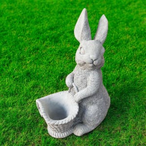 May include: A gray concrete rabbit statue holding a woven basket. The rabbit has long ears and is sitting on a green lawn. The statue is a decorative garden accent.