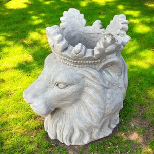 May include: A gray concrete lion head planter with a crown. The planter is set on green grass. The lion's face is detailed with fur and facial features. The crown is ornate and sits atop the lion's head.