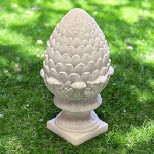 May include: A large, off-white, outdoor garden sculpture shaped like a pinecone. The sculpture has a detailed, textured surface and sits on a square base. The sculpture is placed on a bed of green grass.