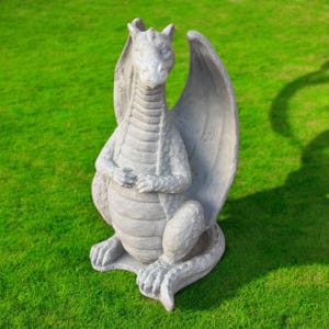 May include: A gray concrete dragon statue with wings, sitting on a green grassy lawn. The dragon has a detailed, scaly texture and is posed with its wings spread out.
