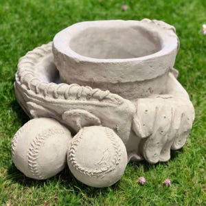 May include: A gray concrete baseball-themed planter with a glove-shaped base, a central pot, and two baseballs. The planter is designed to hold plants and is suitable for outdoor use. The baseballs have detailed stitching.
