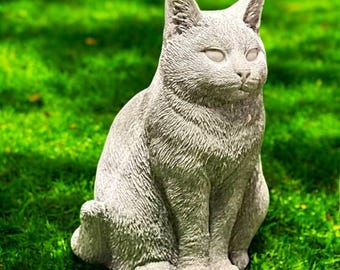 May include: A gray cat statue with a textured surface, sitting on green grass. The cat is in a seated position with its ears perked up. The statue has a detailed fur texture and is a decorative garden ornament.