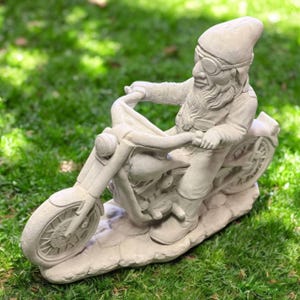 May include: A light gray garden gnome statue riding a motorcycle. The gnome wears goggles and a pointed hat. The motorcycle has detailed wheels and a sculpted engine. The statue is set on a stone-like base, perfect for outdoor decor.