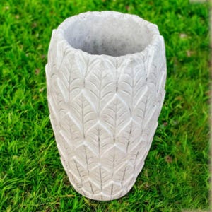 May include: A gray, textured, concrete planter with a leaf pattern. The open top planter is cylindrical and sits on green grass. The leaf pattern is repeated around the entire planter.