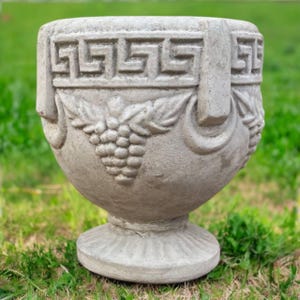 May include: A gray, concrete planter with a pedestal base and two handles. The planter features a Greek key design around the top and grape cluster carvings. The planter is sitting on green grass.