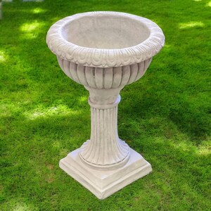 May include: A light gray, ornate pedestal planter with a wide, decorative rim. The planter has a fluted column and a square base. The planter is made of a stone-like material and is set on a green lawn.