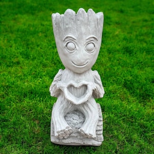 May include: A gray, seated statue of a character with a smiling face and large eyes, holding a heart shape with its hands. The statue has a textured, bark-like appearance and is set against a backdrop of green grass. The statue is a decorative garden ornament.