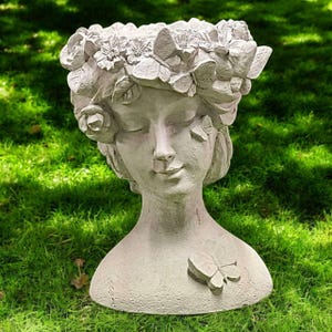 May include: A gray, sculpted head planter with closed eyes, adorned with a floral and butterfly crown. A butterfly is sculpted on the chest. The planter is set against a backdrop of green grass.