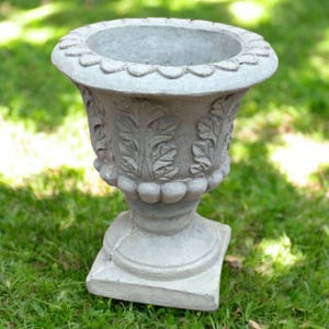 May include: A gray concrete planter with a decorative leaf design and a scalloped rim. The planter is sitting on a bed of green grass.