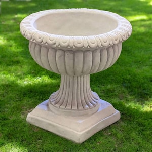 May include: A beige, ornate planter with a wide, scalloped rim. The planter has a fluted body and sits on a square base. The outdoor garden decor is made of a durable material.