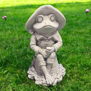 May include: A gray frog statue wearing a hat and a button-down shirt, seated with crossed legs. The frog's hands are clasped, and it is sitting on a textured base. The statue is outdoors on a green lawn.