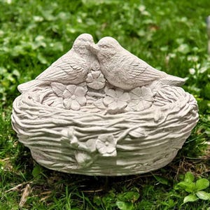 May include: A light gray bird sculpture featuring two birds nestled together in a nest. The nest is intricately detailed with a woven texture and adorned with small flowers. The sculpture is made of a durable material and is suitable for outdoor display.