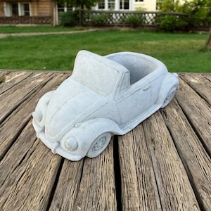 May include: A light gray concrete planter shaped like a vintage convertible car. The planter features a hollow interior, rounded fenders, circular headlights, and a windshield frame, all rendered in a smooth, matte, stone-like material finish.