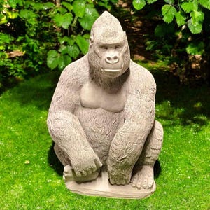 May include: A detailed, gray gorilla statue in a seated position, with its hands resting on its knees. The gorilla has a textured fur appearance and is set against a backdrop of green grass and foliage, suitable for garden decor.