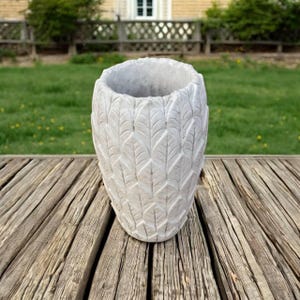 May include: A cylindrical light gray concrete planter featuring a repeating embossed leaf pattern across its entire exterior surface. The vessel has a wide circular opening at the top and a solid base, showcasing a textured finish throughout the design.